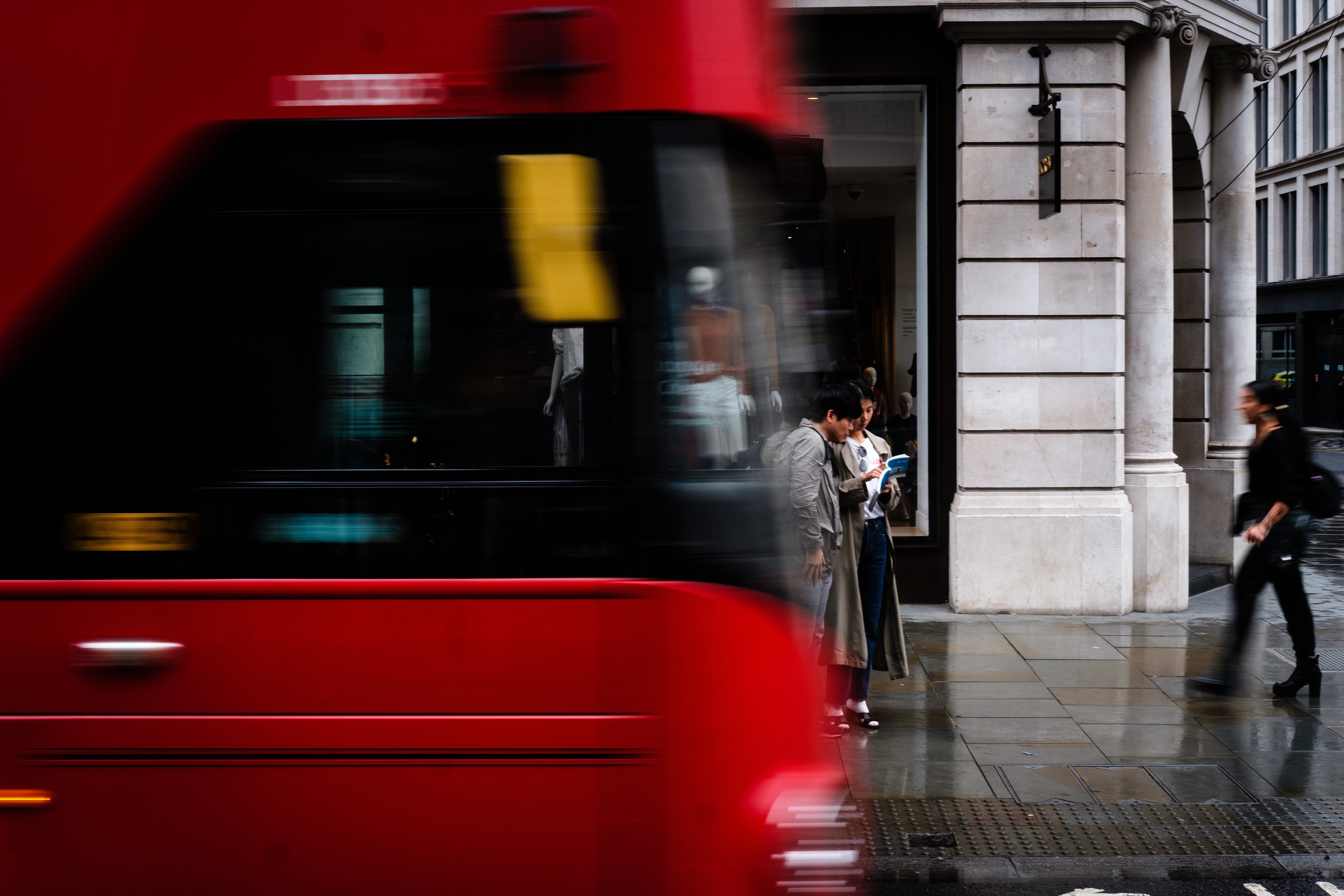 A red London bus blurs past two people reading outside a shopfront on a wet London street.